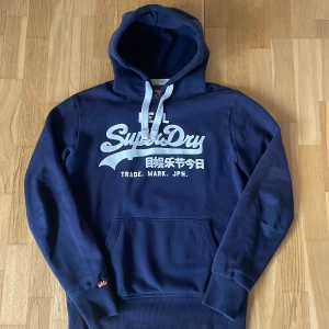 Mörkblå hoodie från Superdry - Säljer en snygg mörkblå hoodie från Superdry med vit text på framsidan. Storleken är M men passar även dig om har storlek L. Tveka inte att höra av dig vid frågor/funderingar!😁💯