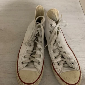 Vita Converse All Star sneakers - Klassiska vita Converse All Star sneakers i hög modell med snörning. Skorna har en röd rand längs sulan och Converse-loggan på sidan. Använt några gånger.