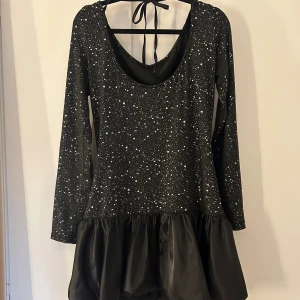 Svart glittrig klänning med volang - Snygg svart klänning med glittrigt mönster och volangdetalj. Den har långa ärmar och en rund halsringning med knytband i nacken. Perfekt för en kväll ute eller en festlig tillställning.