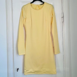 Gul klänning med volangdetalj - Oanvänd Snygg gul klänning med öppen rygg och volangdetalj. Perfekt för en stilren look med en touch av elegans. Klänningen har en enkel och klassisk passform som gör den lätt att bära vid olika tillfällen. 