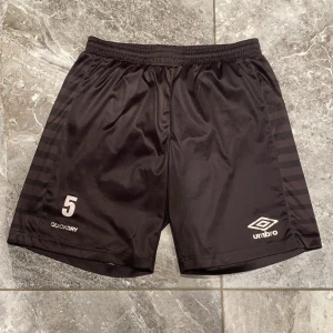 Svarta träningsshorts från Umbro - Snygga svarta träningsshorts från Umbro med elastisk midja och Quickdry-teknologi. Perfekta för fotboll eller annan träning. Diskreta ränder på sidorna och nummer 5 tryckt på framsidan.