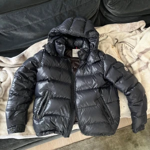 Moncler Maya - Säljer en snygg navy dunjacka från Moncler med huva och dragkedja. Jackan har ett quiltat mönster och Moncler-logga på ärmen. Perfekt för kyliga dagar med sin varma och stilrena design. Storlek 5(L-XL)