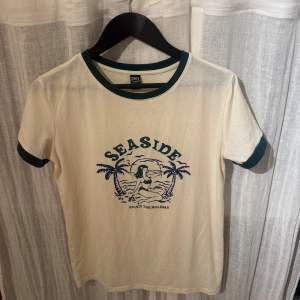 Säljer en vit t-shirt från Dazy med ett blått tryck av en strandscen och texten 'Seaside'. T-shirten har gröna kanter vid hals och ärmslut. Perfekt för en avslappnad stil.