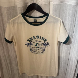 Vit t-shirt med tryck från Dazy - Säljer en vit t-shirt från Dazy med ett blått tryck av en strandscen och texten 'Seaside'. T-shirten har gröna kanter vid hals och ärmslut. Perfekt för en avslappnad stil.