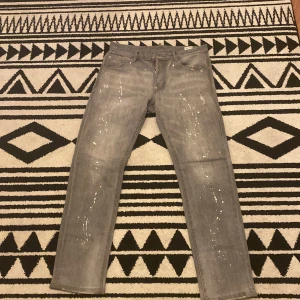 Grå jeansbyxor med färgstänk - Snygga grå jeansbyxor från Dsquared2 med unika färgstänk för en cool look. Byxorna har en straight passform och klassisk femficksdesign. Perfekta för dig som vill sticka ut med stil det är helt nya kom dm vid intresse!