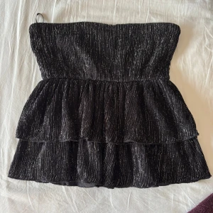 Svart glittrig tubtopp - Snygg svart tubtopp med glittrande detaljer och volanger. Perfekt för en kväll ute eller festliga tillfällen. Toppen har en smickrande passform och är tillverkad i ett glittrigt material som fångar ljuset fint.