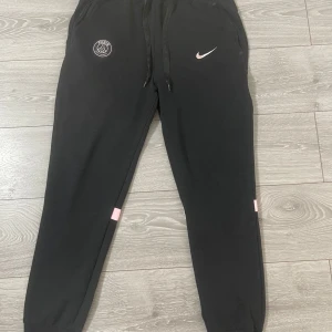 Svarta mjukisbyxor från Nike med PSG-logga - Svarta mjukisbyxor från Nike med PSG-logga och rosa detaljer. Byxorna har snörning i midjan och elastiska muddar vid bensluten. Perfekta för en avslappnad stil.