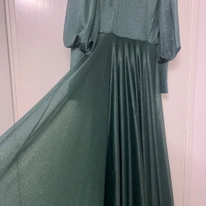 Glittrig grön långklänning - Elegant grön långklänning med glittrande detaljer. Klänningen har långa ärmar och en flytande kjol som ger en vacker silhuett. Perfekt för speciella tillfällen där du vill glänsa.