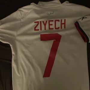 Hakim Ziyech fotbollströja - Säljer en vit fotbollströja från Puma med röd text och nummer 7 på ryggen. Tröjan har korta ärmar och ett snyggt mönster på framsidan med ett emblem i mitten. Perfekt för fotbollsfans! ⚽