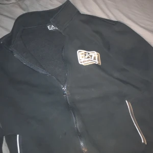 Svart jacka från EA7 Emporio Armani - Snygg svart jacka från EA7 Emporio Armani med dragkedja och broderad logga på bröstet. Jackan har en stilren design med långa ärmar och praktiska fickor med dragkedja. Perfekt för en casual look.