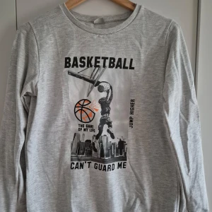 Grå sweatshirt med baskettryck - Säljer en grå sweatshirt med ett coolt baskettryck på framsidan. Tröjan har långa ärmar och en rund halsringning. Perfekt för sportiga tillfällen eller avslappnade dagar. 🏀