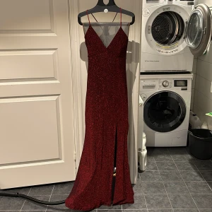 Vinröd glittrig långklänning - Säljer en elegant vinröd glittrig långklänning med smala axelband och v-ringning. Klänningen har en slits framtill och en snygg snörning i ryggen. Använd 1 gång och är i väldigt stretchigt tyg