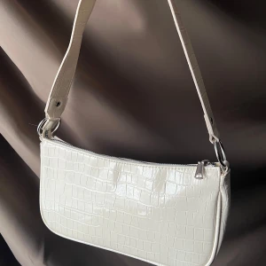 Vit axelväska med krokodilmönster - Snygg vit axelväska med krokodilmönster. Väskan har en dragkedja upptill för säker förvaring. Perfekt för att ge en stilren touch till din outfit.