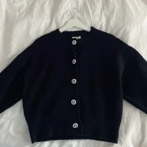 Svart kofta med pärlknappar från H&M - Säljer en stilren svart kofta från H&M med dekorativa pärlknappar framtill. Koftan har en klassisk design med långa ärmar och rund halsringning. Perfekt för att lägga till en touch av elegans till din outfit.