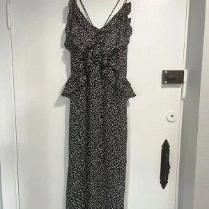 Svart långklänning med prickigt mönster - Elegant svart långklänning med vita prickar och volangdetaljer. Klänningen har en öppen rygg och smala axelband som knyts i nacken. Perfekt för en stilfull look.