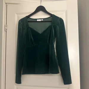 Grön sammetstopp från Vila - Snygg grön sammetstopp från Vila med långa ärmar och en elegant v-ringning. Perfekt för att ge en lyxig känsla till din outfit. Toppen har en figursydd passform som framhäver silhuetten.