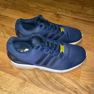 Blå Adidas ZX Flux sneakers - Säljer ett par blå Adidas ZX Flux sneakers med klassiska tre ränder på sidan. Skorna har snörning och en vit sula med Torsion-system för extra stöd. Perfekta för en sportig look.