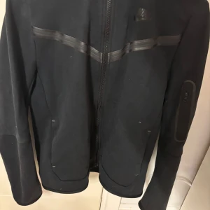Svart jacka från Nike - Säljer en svart jacka från Nike med dragkedja och huva. Jackan har en stilren design med diskreta detaljer och en ficka med dragkedja på ärmen. Perfekt för en sportig look. Priset kan diskuteras Nike teech hoodie haft i ett halvår köpt från Nike hemsidan.Det är två fickor på båda av sidorna inga fläckar eller hål i tröjan.