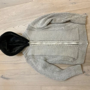 Grå stickad cardigan med pälsluva - Säljer en grå stickad cardigan med dragkedja och mjukt foder i luvan. Perfekt för kyliga dagar med sin ribbade design och långa ärmar. En mysig och stilren tröja för alla tillfällen. Passar M storlek M!