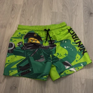 Gröna Ninjago shorts från H&M - Coola gröna badshorts med Ninjago-tryck från H&M. Perfekta för sommarens äventyr med elastisk midja och snörning för bästa passform. Tillverkade i lätt och bekvämt material.