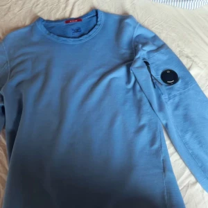 Blå långärmad tröja från C.P. Company - Säljer en snygg blå långärmad tröja från C.P. Company. Tröjan har en unik detalj med en rund logotyp på ärmen och är tillverkad i ett mjukt material. Perfekt för en stilren look. Pris kan diskuteras!