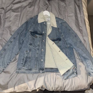 Vinter Jeansjacka - Säljer en snygg blå jeansjacka med vit teddyfoder. Jackan har en klassisk krage och knappar framtill. Perfekt för kyligare dagar med sin varma insida. Två bröstfickor med lock och två sidofickor ger extra stil och funktion. Jackan har storlek XL men sitter enligt mig som storlek Large. 