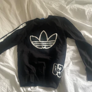 Svart långärmad tröja från Adidas - Säljer en svart långärmad tröja från Adidas med det klassiska trefoil-logotypen i vitt på framsidan. Tröjan har en sportig look med vita ränder längs ärmarna och siffran '03' tryckt på nedre delen. Perfekt för en casual stil eller träning.