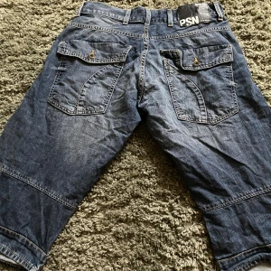 Blå jeansshorts - Säljer ett par blå jeansshorts från PSN med knappar och fickor bak. De har en loose passform och är perfekta för en avslappnad stil. Shortsen har en klassisk jeanslook med slitningar och är knälånga. Passar både män och kvinnor!