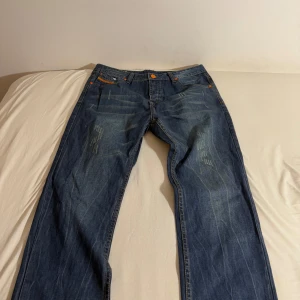 Blå jeans från Diesel X Adidas - MADE IN ITALY. Snygga blå jeans från Diesel med orange detaljer och en klassisk femficksdesign. Jeansen har en straight passform och är tillverkade i 100% cotton denim. Perfekta för en avslappnad stil. Notera en mindre defekt på adidas loggan på baksidan. (W: ca 45 cm diameter, L32)