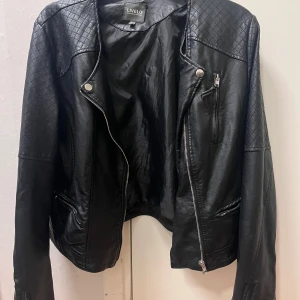 Svart skinnjacka från Livelo - Snygg svart skinnjacka från Livelo med quiltade detaljer på axlar och ärmar. Jackan har en asymmetrisk dragkedja och flera fickor med dragkedjor. Perfekt för en cool och stilren look.