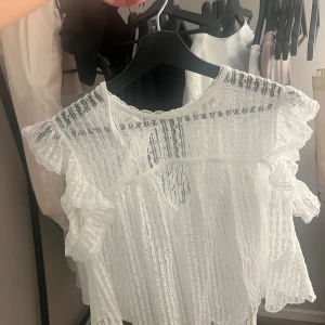 Vit spetsblus med volanger - Säljer en söt vit blus i spets med volangdetaljer på ärmarna. Blusen har en knytning i nacken och är perfekt för en romantisk look. Den är kortärmad och har en luftig design.