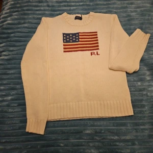 Vit stickad tröja från Ralph Lauren - Säljer en vit stickad tröja från Ralph Lauren med ett amerikanskt flaggmotiv och initialerna 'RL' på framsidan. Tröjan har långa ärmar och en rund halsringning. Perfekt för en stilren och klassisk look.