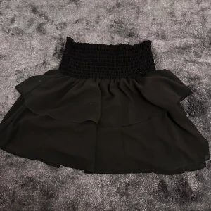 Svart kjol från Chelsea - Snygg svart volangkjol från Chelsea med en elastisk midja som ger en bekväm passform. Kjolen har en volangdetalj som ger en extra touch av stil. Perfekt för att skapa en chic look.