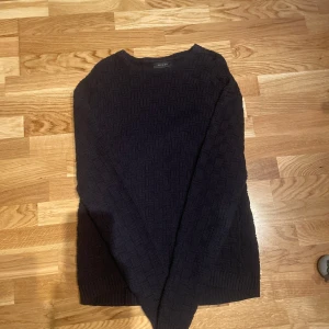 Svart stickad tröja från Religion - Säljer en stilren svart stickad tröja från Religion. Tröjan har ett diskret mönster och är perfekt för kyligare dagar. Den har långa ärmar och en klassisk passform. Perfekt för dig som gillar en minimalistisk stil.