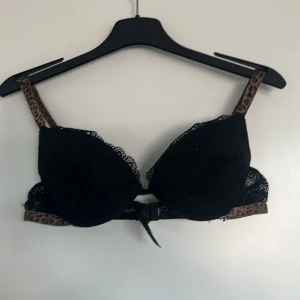 Svart spets-bh med leoparddetaljer - Snygg svart bh med spets och leopardmönstrade axelband. Den har en dekorativ rosett framtill och ger en elegant look. Perfekt för att lägga till lite extra stil i garderoben.
