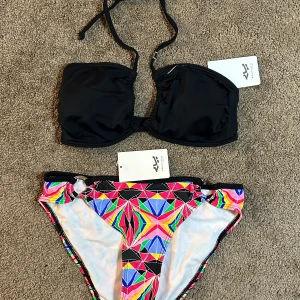 NY Svart och mönstrad bikini från Röhnisch - Trendig bikini från Röhnisch med en svart bandeau-topp och färgglatt mönstrade trosor. Toppen har knytband och en subtil rynkdetalj framtill. Trosorna har ett geometriskt mönster i neonfärger som verkligen sticker ut. Perfekt för en dag på stranden! Nypris ca 800kr