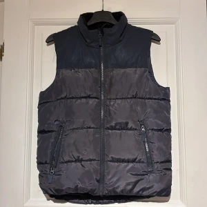 Mörkblå dunväst från Wave - Snygg och praktisk mörkblå dunväst från Wave med dragkedja framtill. Västen har två fickor med dragkedjor och en hög krage för extra värme. Perfekt för kyliga dagar.