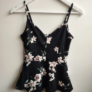 Svart blommig linnetopp - Snygg svart linnetopp med blommigt mönster i rosa och vitt. Toppen har smala axelband och en lätt utsvängd nederdel. Perfekt för varma dagar eller sommarens fester.