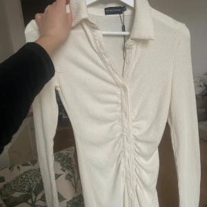 Vit ribbad klänning från PrettyLittleThing - Snygg vit ribbad klänning från PrettyLittleThing med långa ärmar och knappar framtill. Klänningen har en stilren design med en lätt rynkad effekt längs knäppningen. Aldrig använd 😊
