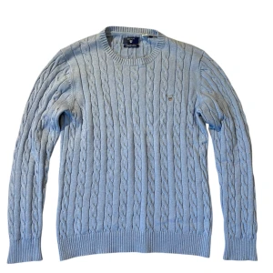 Ljusblå kabelstickad tröja från GANT - Säljer en stilren ljusblå kabelstickad tröja från GANT i premium bomull. Tröjan har ett broderat emblem på bröstet och ribbade muddar vid ärmslut och nederkant. Perfekt för en klassisk look.