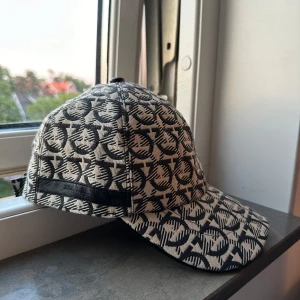 Ferragamo keps - Snygg keps med ett unikt mönster i svart och beige. Kepsen har en justerbar rem baktill för perfekt passform. Perfekt för att ge din outfit en stilfull touch.