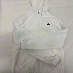 Vit vindjacka från Lacoste - Säljer en stilren vit vindjacka från Lacoste med huva och dragkedja. Jackan har det klassiska Lacoste-logotypen på bröstet och är perfekt för en sportig look. Den är långärmad och har elastiska muddar för en bekväm passform.