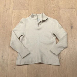 Beige merinoulltröja från These Glory Days - Säljer en stilren beige tröja i merinoull från These Glory Days. Tröjan har en dragkedja vid halsen och långa ärmar, perfekt för kyligare dagar. Den är mjuk och bekväm, vilket gör den till ett utmärkt val för både vardag och mer uppklädda tillfällen. Den är dock lite mindre än s då den har krympt i tvätten.