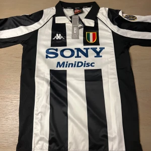Svartvit randig fotbollströja från Kappa - Klassisk svartvit randig fotbollströja från Kappa med SONY MiniDisc-logga på framsidan. Tröjan har korta ärmar och ett emblem på bröstet. På baksidan finns namnet och numret 'Zidane 21'. Perfekt för fotbollsfans och samlare!