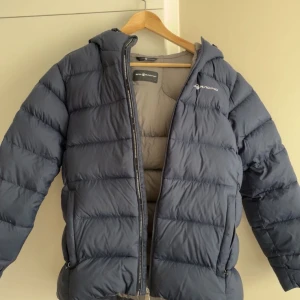 Mörkblå dunjacka från sail Racing - Säljer en mörkblå dunjacka från SAIL RACING  med huva och dragkedja. Jackan är perfekt för kyliga dagar och har en stilren design. Den är vadderad för extra värme och komfort. (EGNA BILDER PRIVAT) 