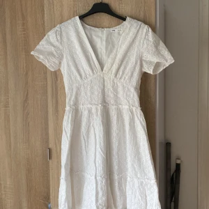 Studentkläning: Vit klänning med broderad spets - En fin studentkläning med v-ringning och korta ärmar. Klänningen har ett vackert broderat spetsmönster och en dragkedja i ryggen. Perfekt för sommarens alla tillfällen. Använd en gång under studenten är i storlek 38 men skulle säga att den passar storlek 36 köpt för 600 
