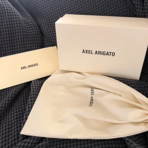 Svarta sneakers från Axel Arigato - Säljer ett par svarta sneakers från Axel Arigato i bra skick. Skorna har en vit sula och en snygg fågeldetalj på sidan. Perfekta för vardagsbruk och ger en stilren look. De kommer med originalförpackning och dustbag.