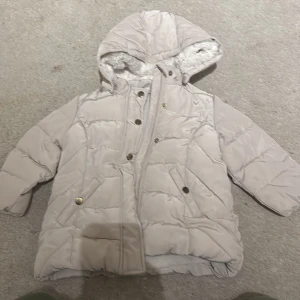 Beige dunjacka med huva - Mysig beige dunjacka med fluffigt foder och huva. Perfekt för kalla vinterdagar. Jackan har knappar och dragkedja framtill för extra värme. Den är i mycket bra skick och har långa ärmar. Passar perfekt för vinterns alla äventyr! FÖR BABYIS!!!