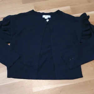 Marinblå trendig kofta från gina tricot! - Säljer en marinblå trendig snygg kofta med en silverstjärn-knapp och volanger vid axlarna/ärmarna. Säljer för att: inte kommit till användning+för liten för mig. Bra skick, använd väldigt fåtal gånger. Nästintill nyskick. PRIS KAN DISKUTERAS!💙
