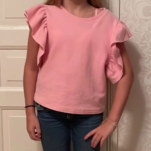 Rosa topp med volangärmar - Säljer en söt rosa topp med volangärmar. Perfekt för vår och sommar! Toppen har en normal passform och är gjord i ett mjukt material som känns skönt mot huden. Den är enkel men ändå stilfull, perfekt för både vardag och festliga tillfällen.Är ett måste i garderoben 